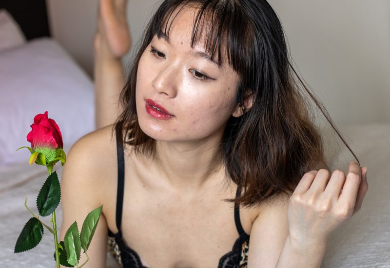 A confident woman holds a single rose close to her chest during a boudoir photoshoot, softly lit against a moody backdrop.